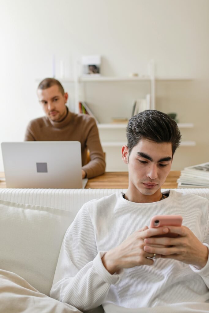 A relaxed young man using a smartphone in a modern home office setting with a laptop.